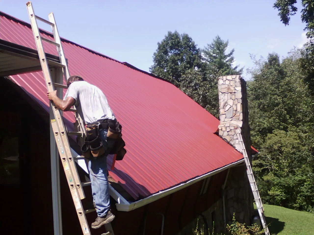 Roofer on a ladder beside a red metal roof and stone chimney — Soffit & Fascia Repair by Roofing Brothers in Taylorstown